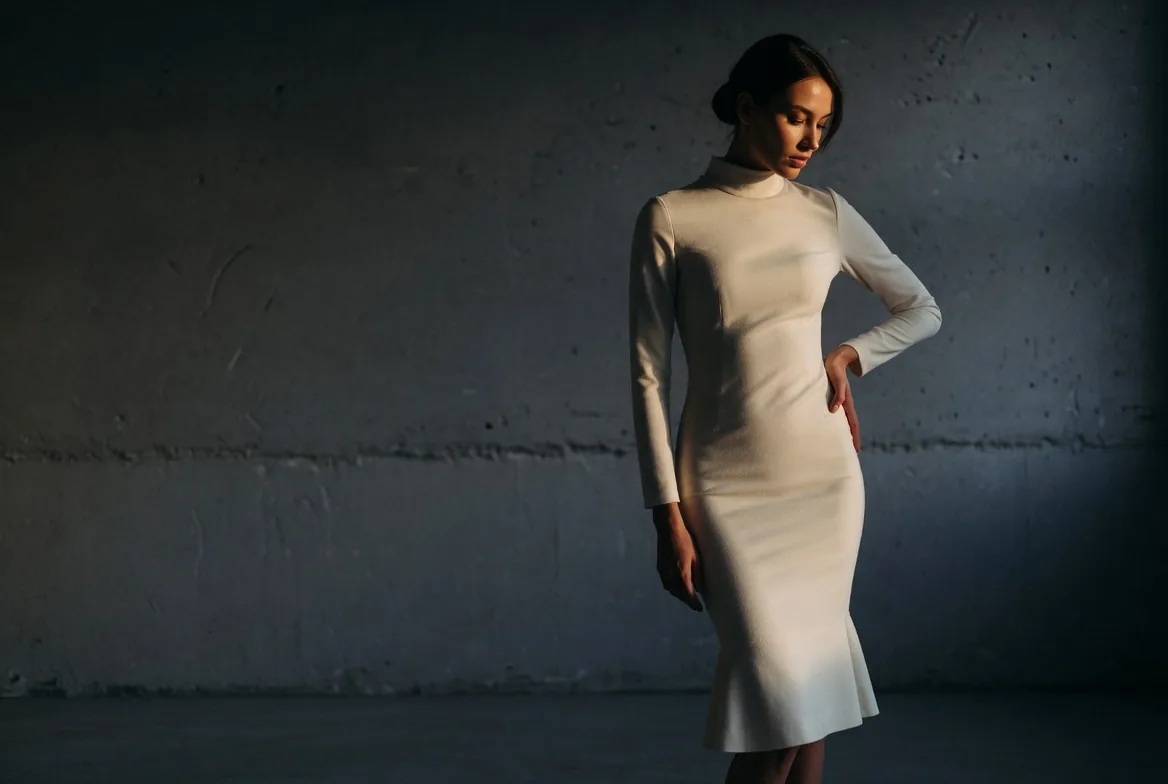 Editorial campaign image: a woman in an ivory fitted dress in a low-lit concrete studio